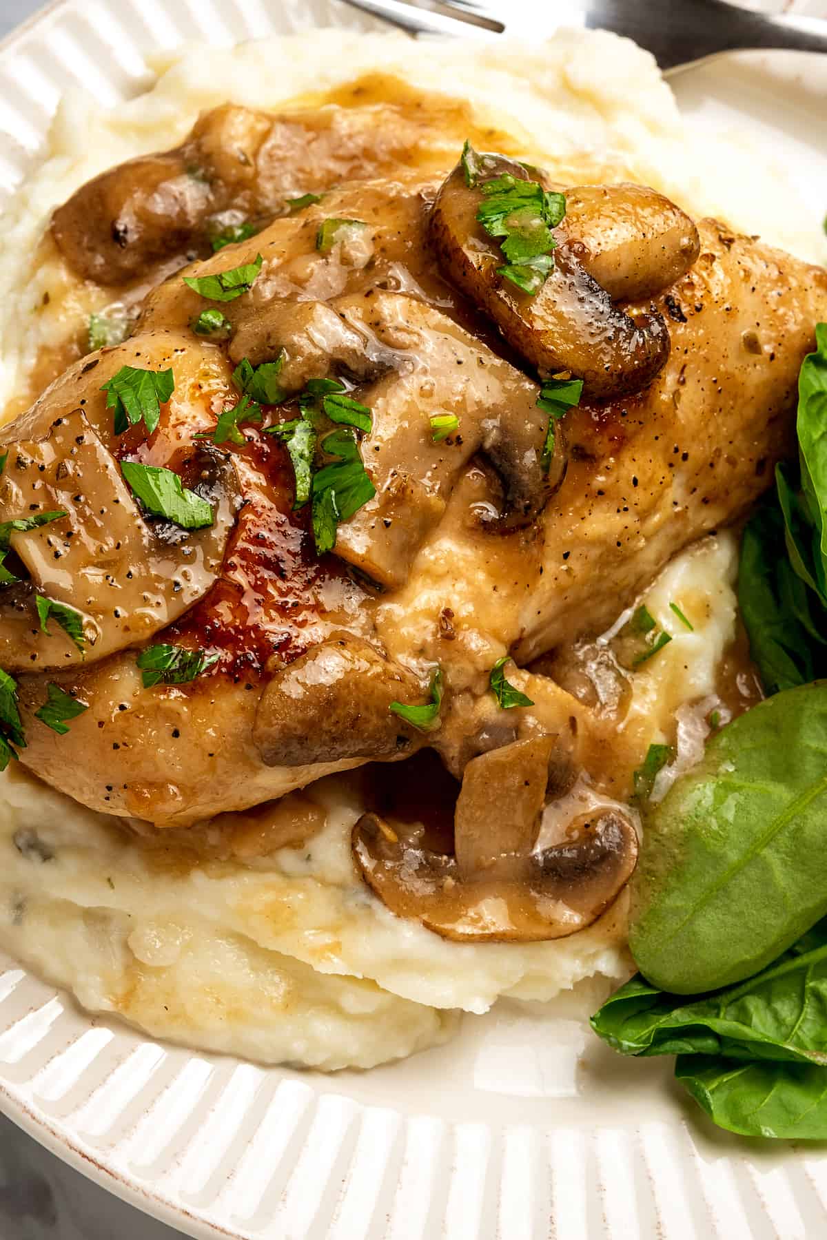 Close up top view of chicken marsala served over a bed of mashed potatoes with a side of greens on a plate.