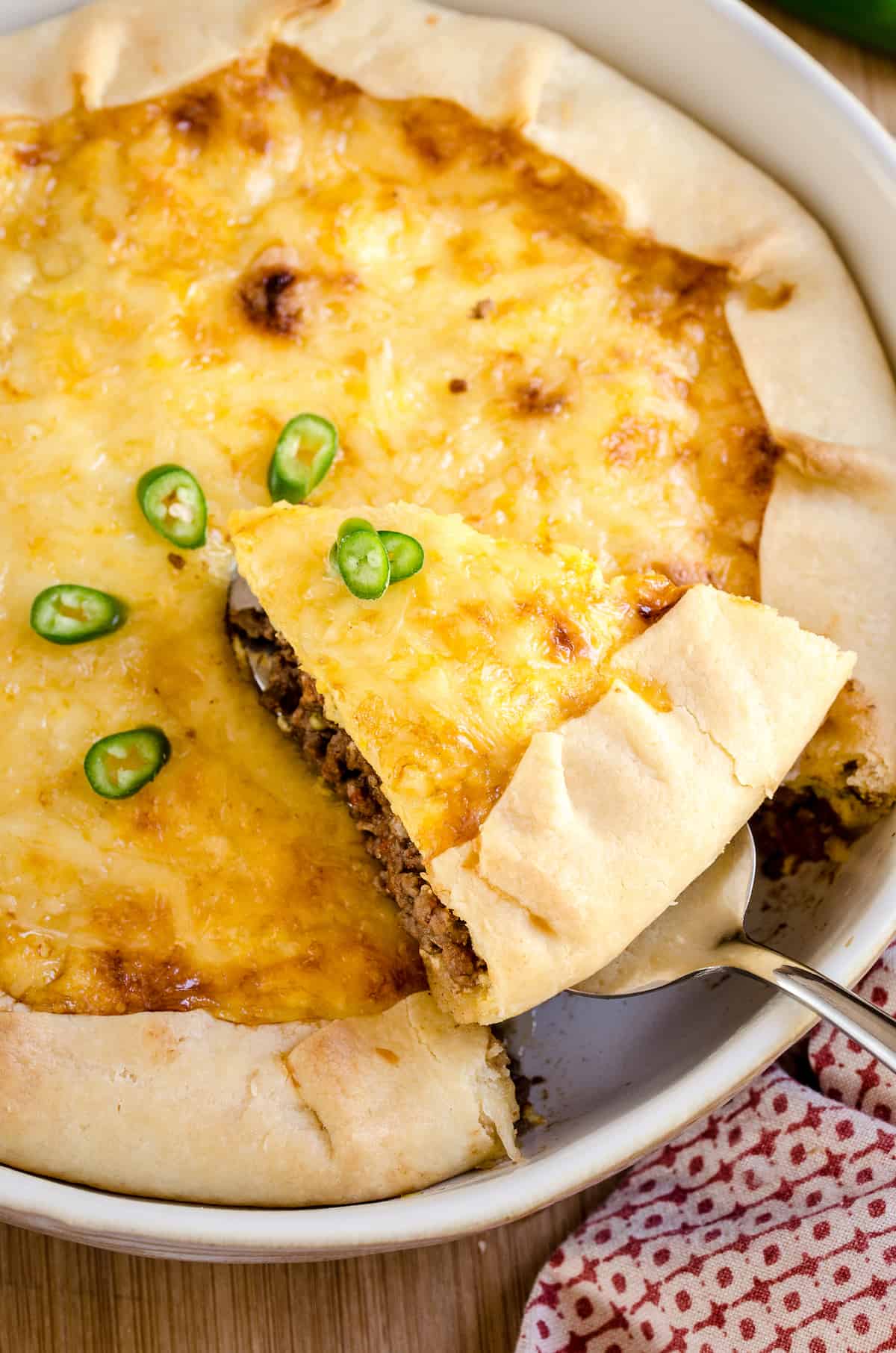 A slice of cheesy quiche topped with green chilies is removed from a fully baked quiche.