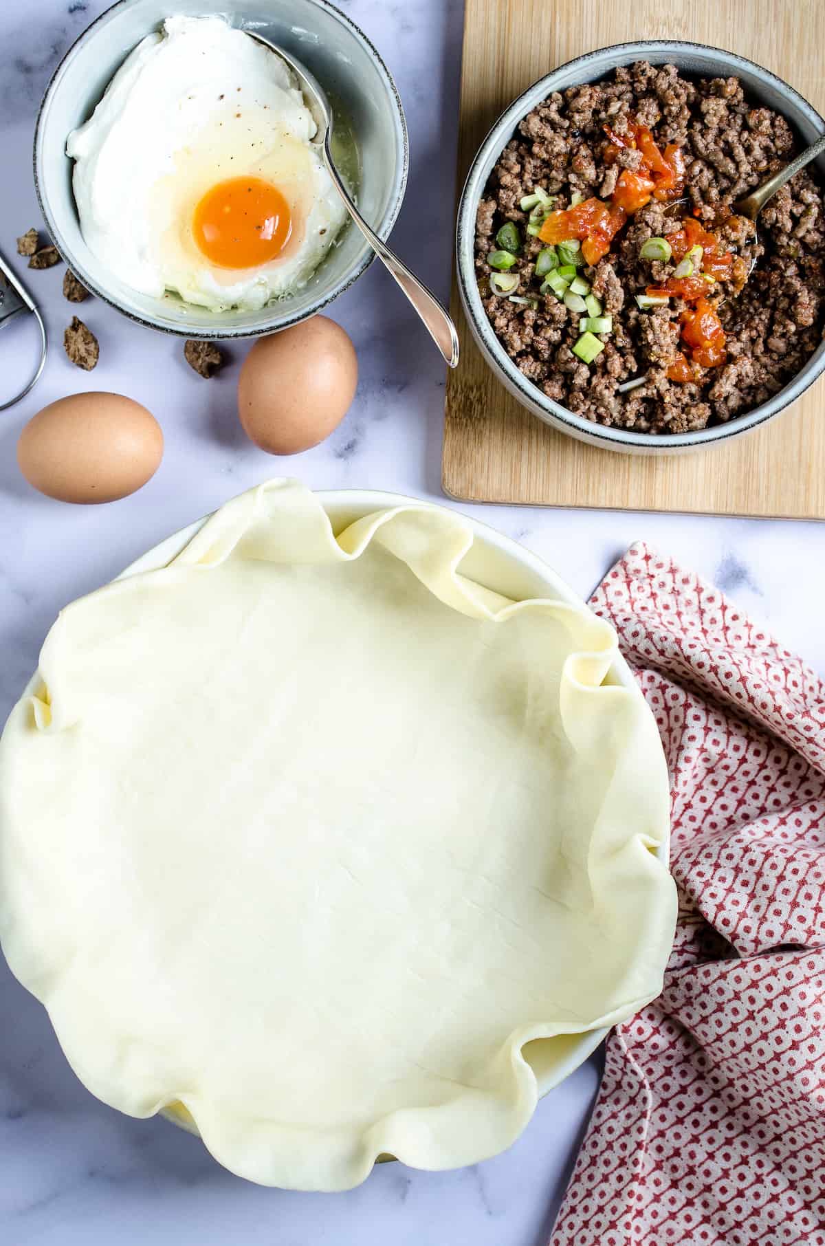 A flaky pie crust lines a prepared baking dish, surrounded by the ingredients for this Tex-Mex Cheesy Beef Quiche recipe.