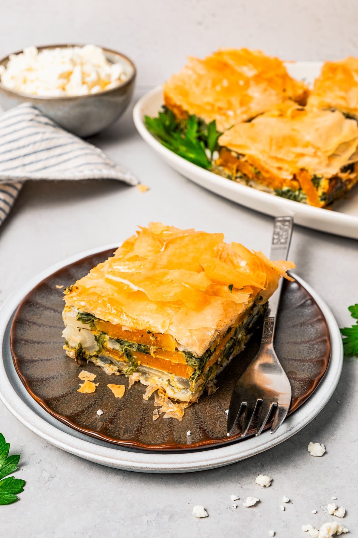 A slice of butternut squash and spinach pie on a plate next to a fork, with additional slices of pie on a platter in the background.