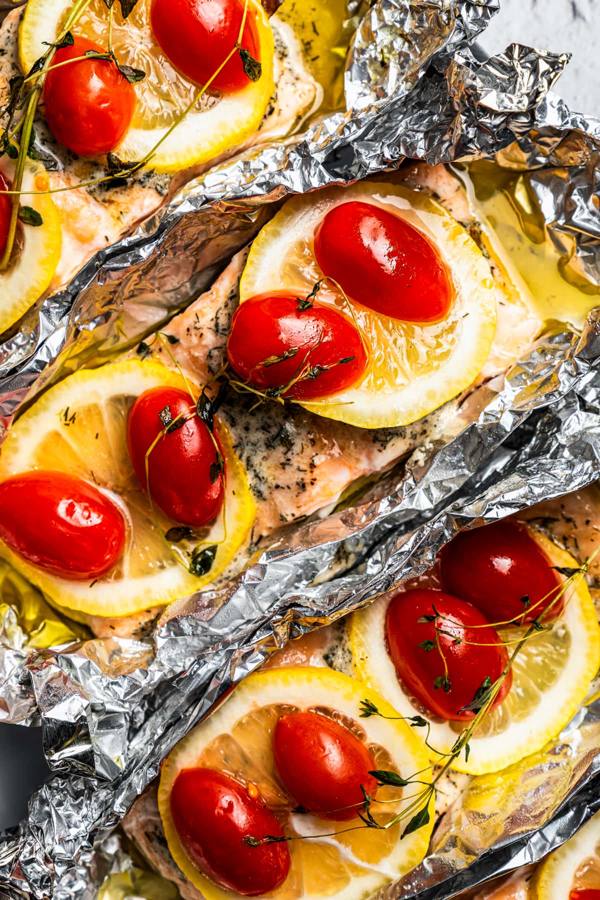 Close up of baked lemon pepper salmon filets topped with lemon slices, tomatoes, and thyme inside unwrapped foil packets.