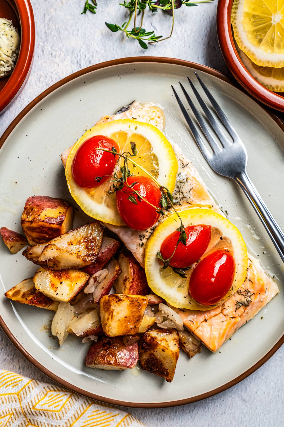 Overhead view of lemon pepper salmon topped with lemon slices, tomatoes, and thyme on a plate next to crispy potatoes and a fork.