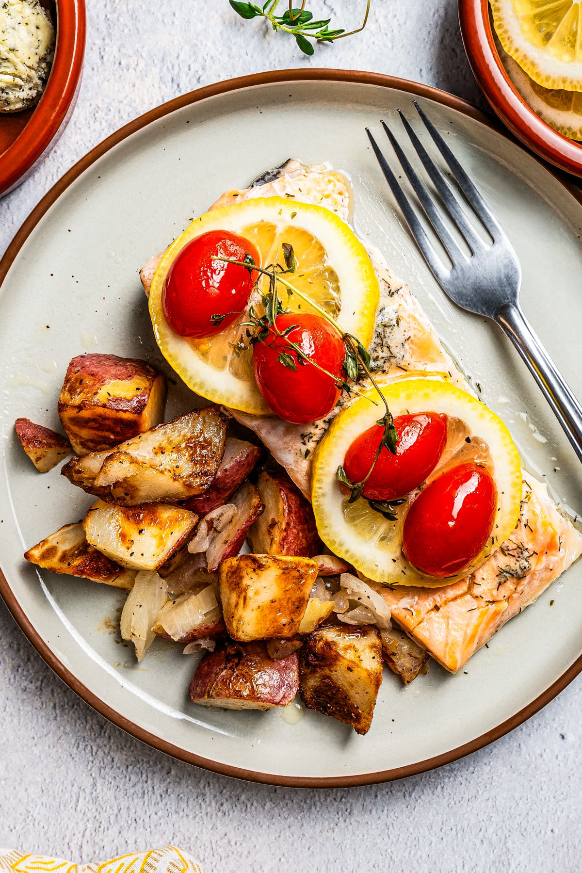 Overhead close up view of lemon pepper salmon topped with lemon slices, tomatoes, and thyme on a plate next to crispy potatoes and a fork.