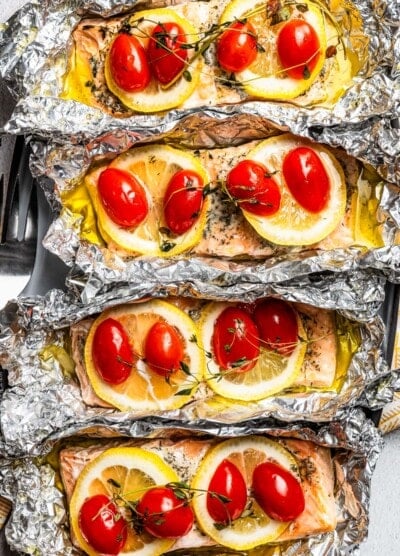 Overhead, close-up view of baked lemon pepper salmon fillets topped with lemon slices, tomatoes, and thyme inside unwrapped foil packets.