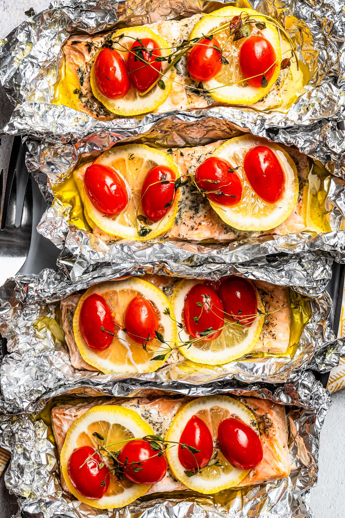 Overhead, close-up view of baked lemon pepper salmon fillets topped with lemon slices, tomatoes, and thyme inside unwrapped foil packets.