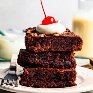 A stack of three 2-ingredient brownies topped with a dollop of whipped cream and a maraschino cherry.