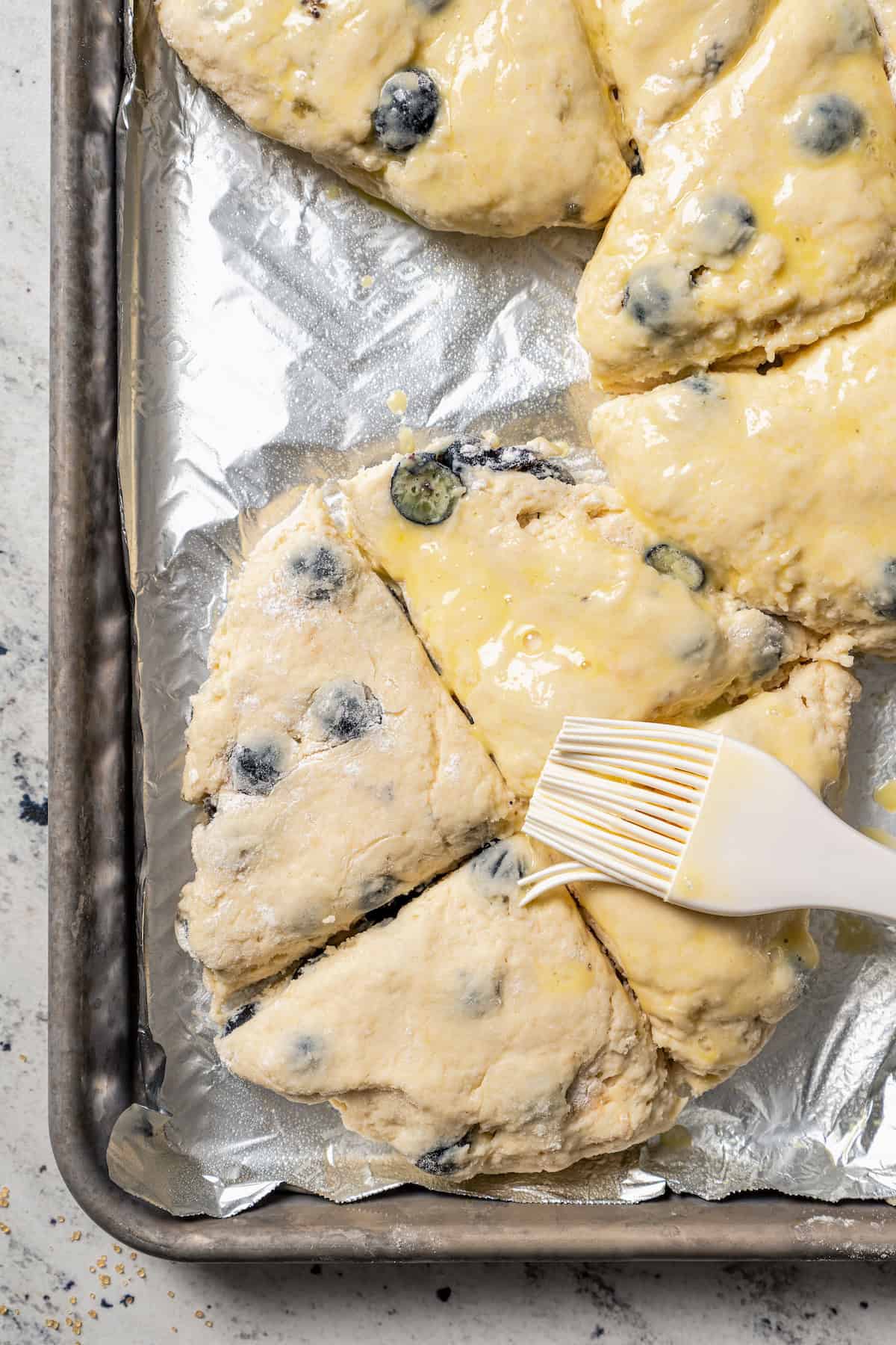 A rubber basting brush brushing butter over quartered blueberry scone dough.