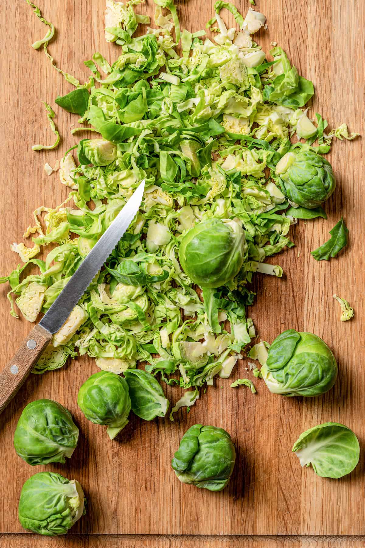 Overhead view of Brussels sprouts shavings next to whole Brussels sprouts on a cutting board, with a sharp knife.