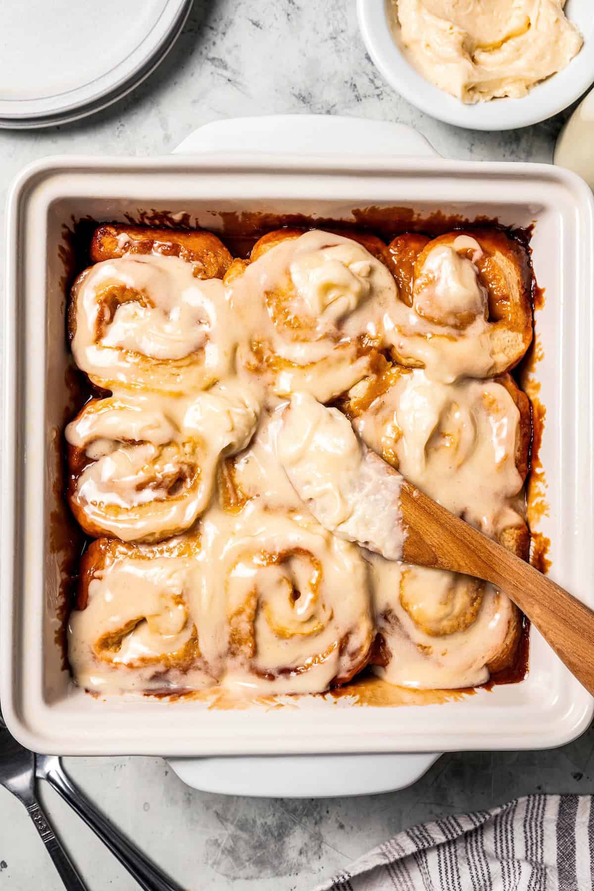 These sweet and gooey homemade caramel rolls require no yeast, rise time, or kneading. They're easy, quick, and best of all, delicious! Overhead view of frosted caramel rolls with a wooden spatula in a baking dish.