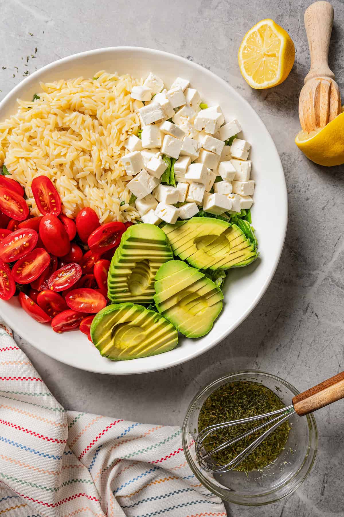 Overhead view of sliced avocado, tomatoes, feta cheese, and orzo pasta arranged in a large bowl next to a mason jar of dressing with a whisk.