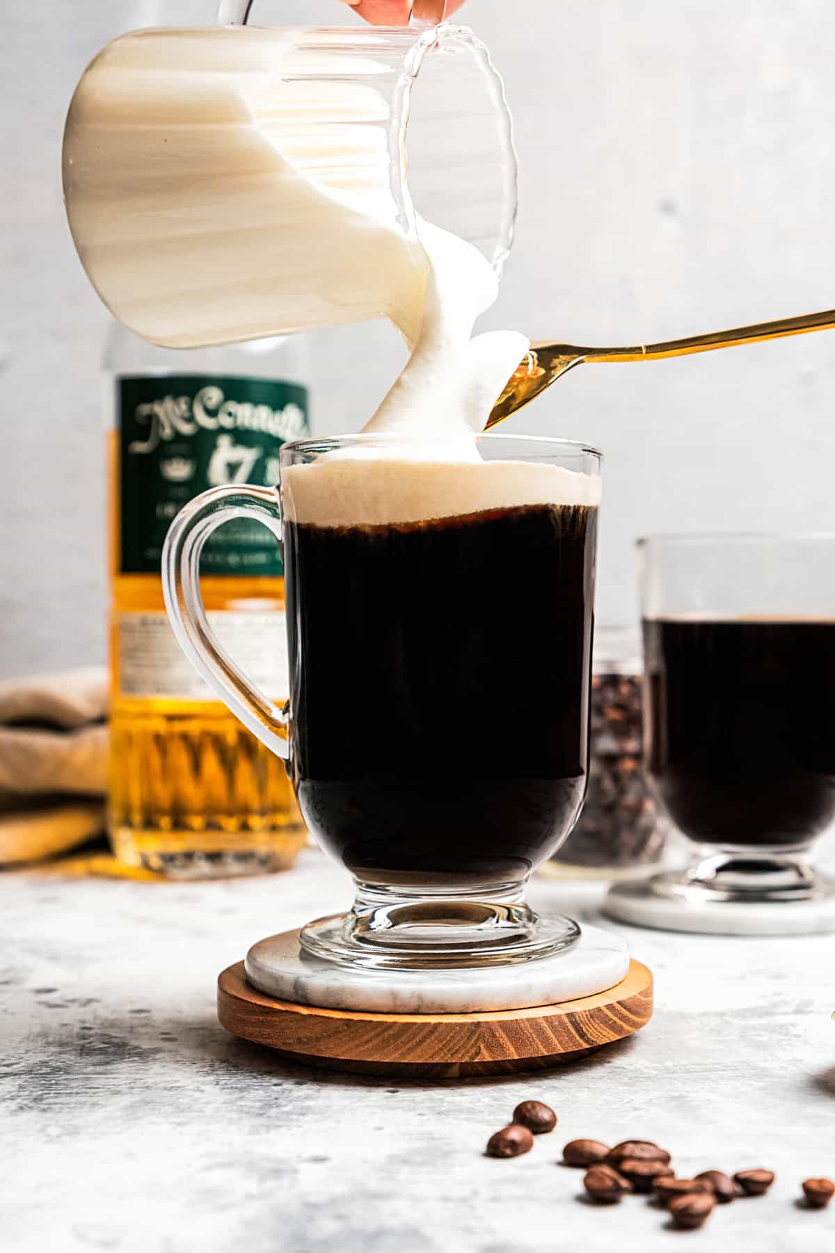 Frothed milk is being poured over the top of the coffee in a glass mug, with a second coffee and a bottle of whiskey in the background.