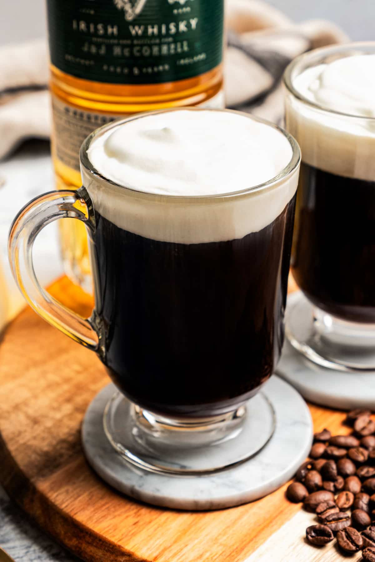 An Irish coffee in a glass mug next to another cup of coffee with coffee beans scattered on a wooden tray, with a bottle of whiskey in the background.