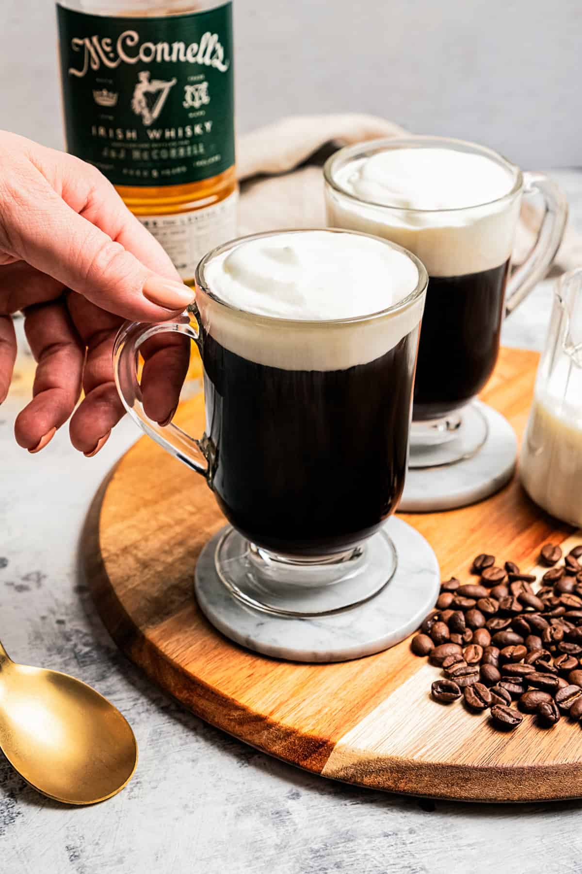 A hand reaching for a mug of Irish coffee next to coffee beans scattered on a wooden tray, with a second mug and a bottle of whiskey in the background.