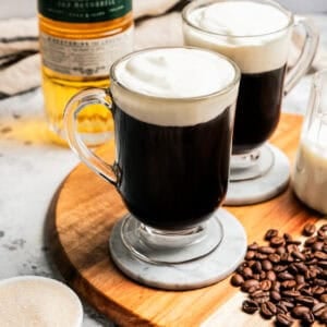 Two Irish coffees in glass mugs next to coffee beans scattered on a wooden tray, with a glass creamer with milk and a bottle of whiskey in the background.