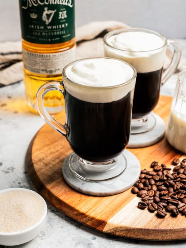 Two Irish coffees in glass mugs next to coffee beans scattered on a wooden tray, with a glass creamer with milk and a bottle of whiskey in the background.