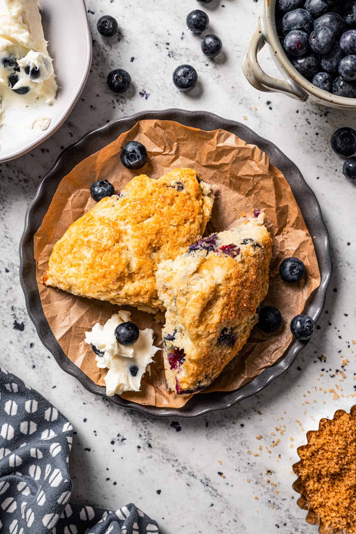 Overhead view of two blueberry scones on a parchment-lined plate next to a dollop of blueberry cream cheese, next to a bowl of blueberries.