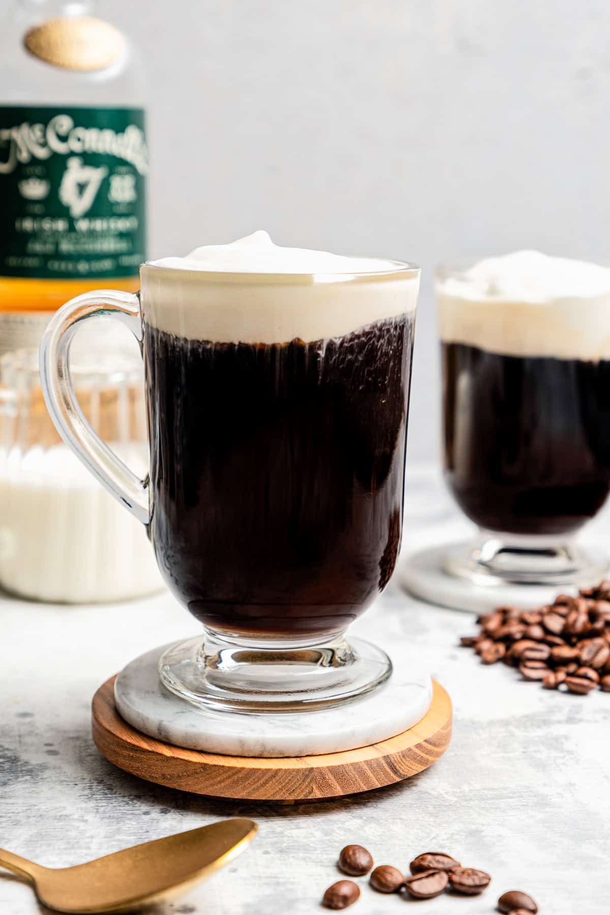 Two mugs of Irish coffee set on coasters with a bottle of whiskey placed in the background.