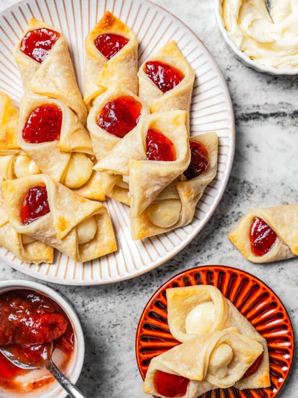 Overhead view of a platter of kolacky cookies next to two cookies on a small red plate and a bowl of strawberry filling.