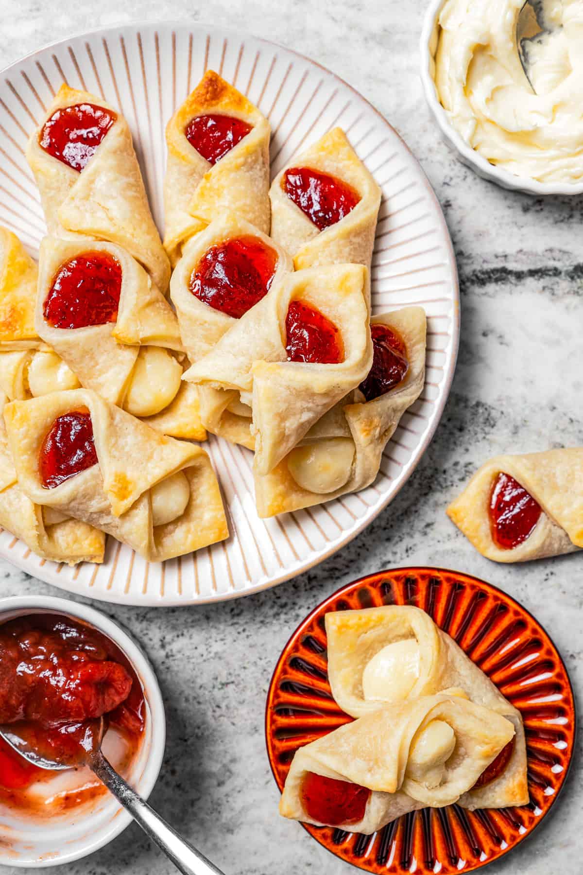 Overhead view of a platter of kolacky cookies next to two cookies on a small red plate and a bowl of strawberry filling.