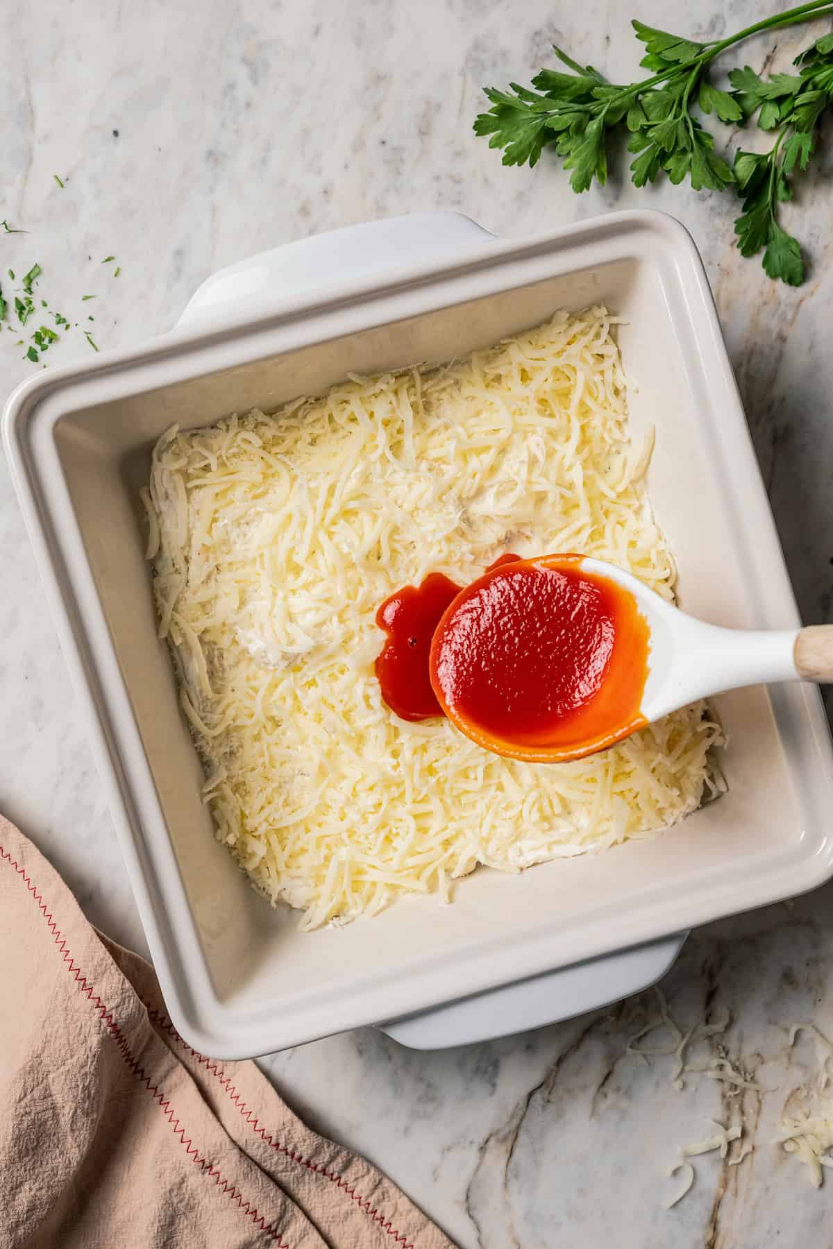 Sauce being added over top a layer of shredded cheese in a baking dish.