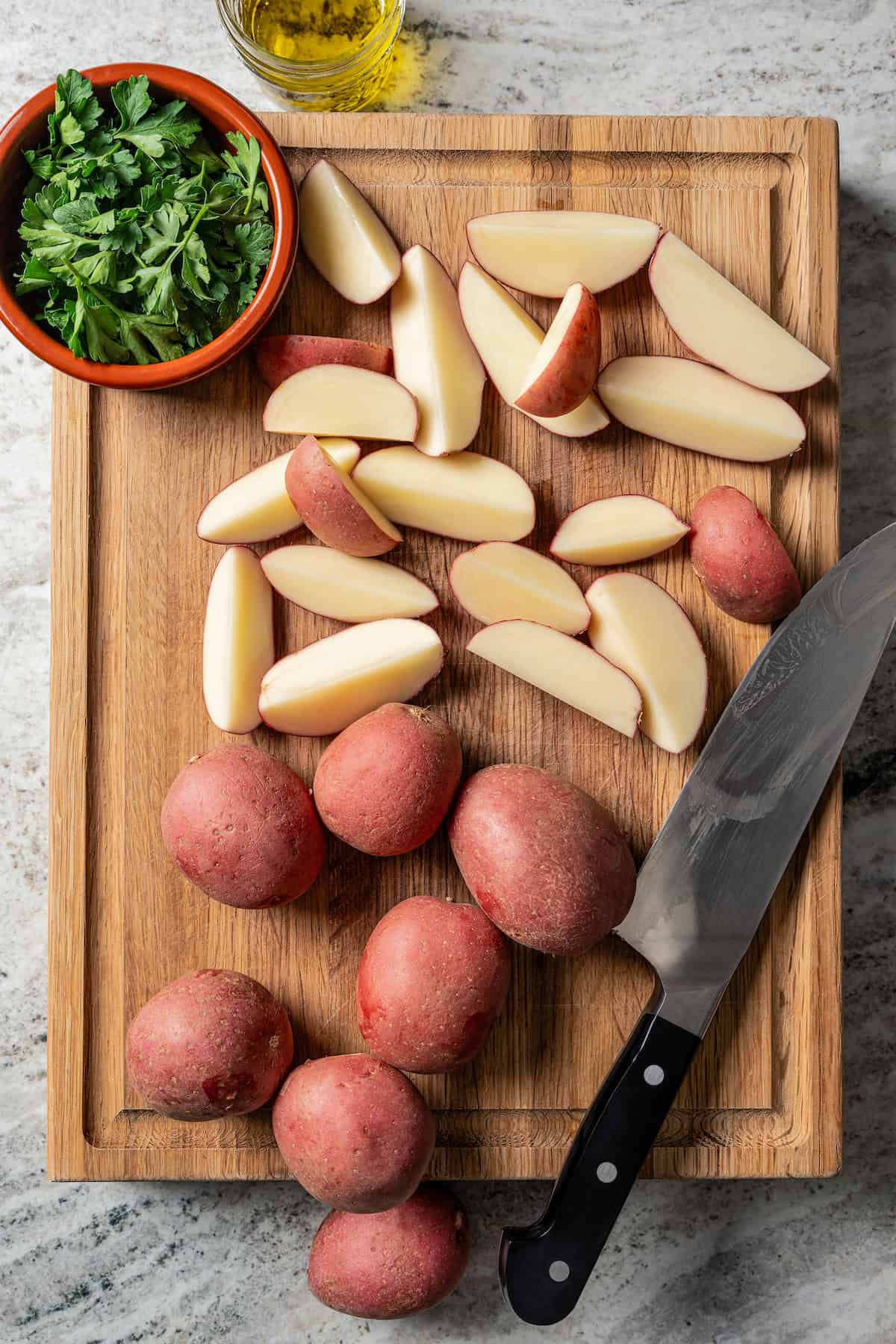 Overhead view of red potatoes partially cut into wedges on a wooden cutting board, next to a sharp knife and a bowl of parsley.