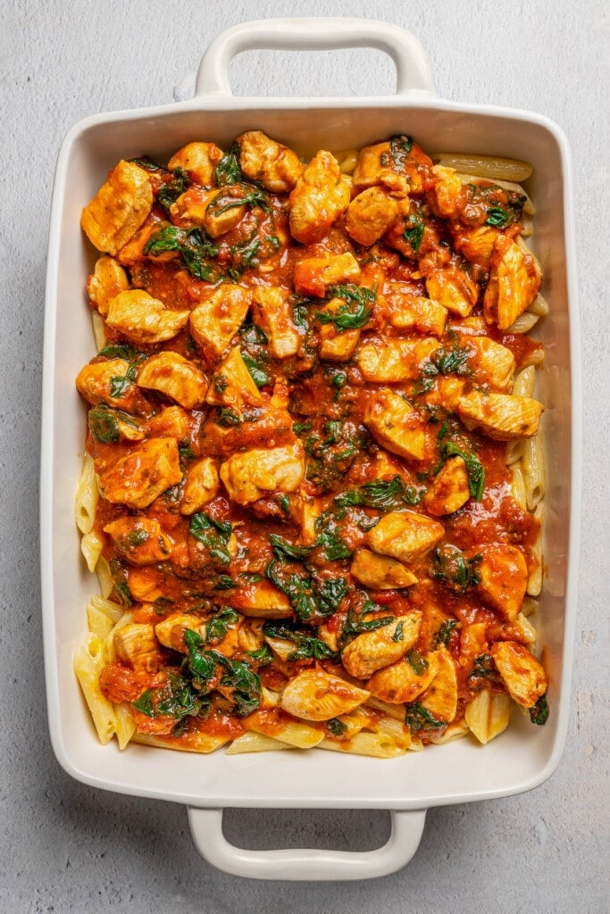 Chicken pieces in tomato sauce layered over top cheesy penne pasta in a casserole dish.