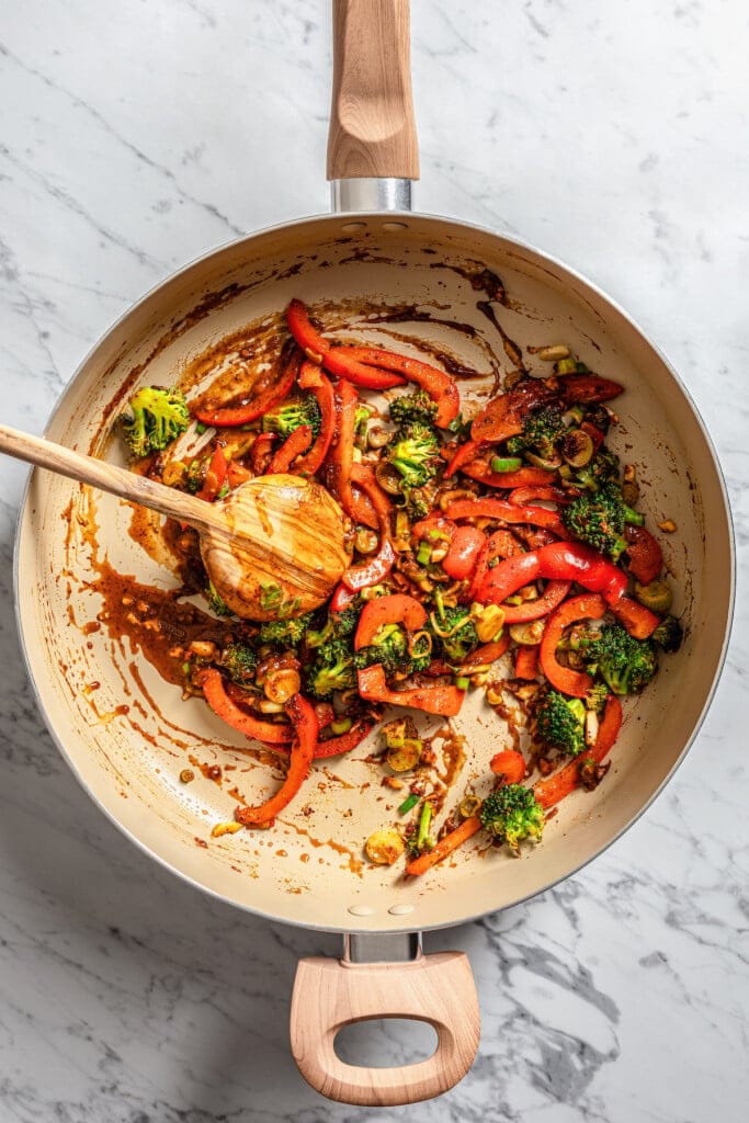 Stir-fried vegetables combined with General Tso's sauce in a large pan with a wooden spoon.