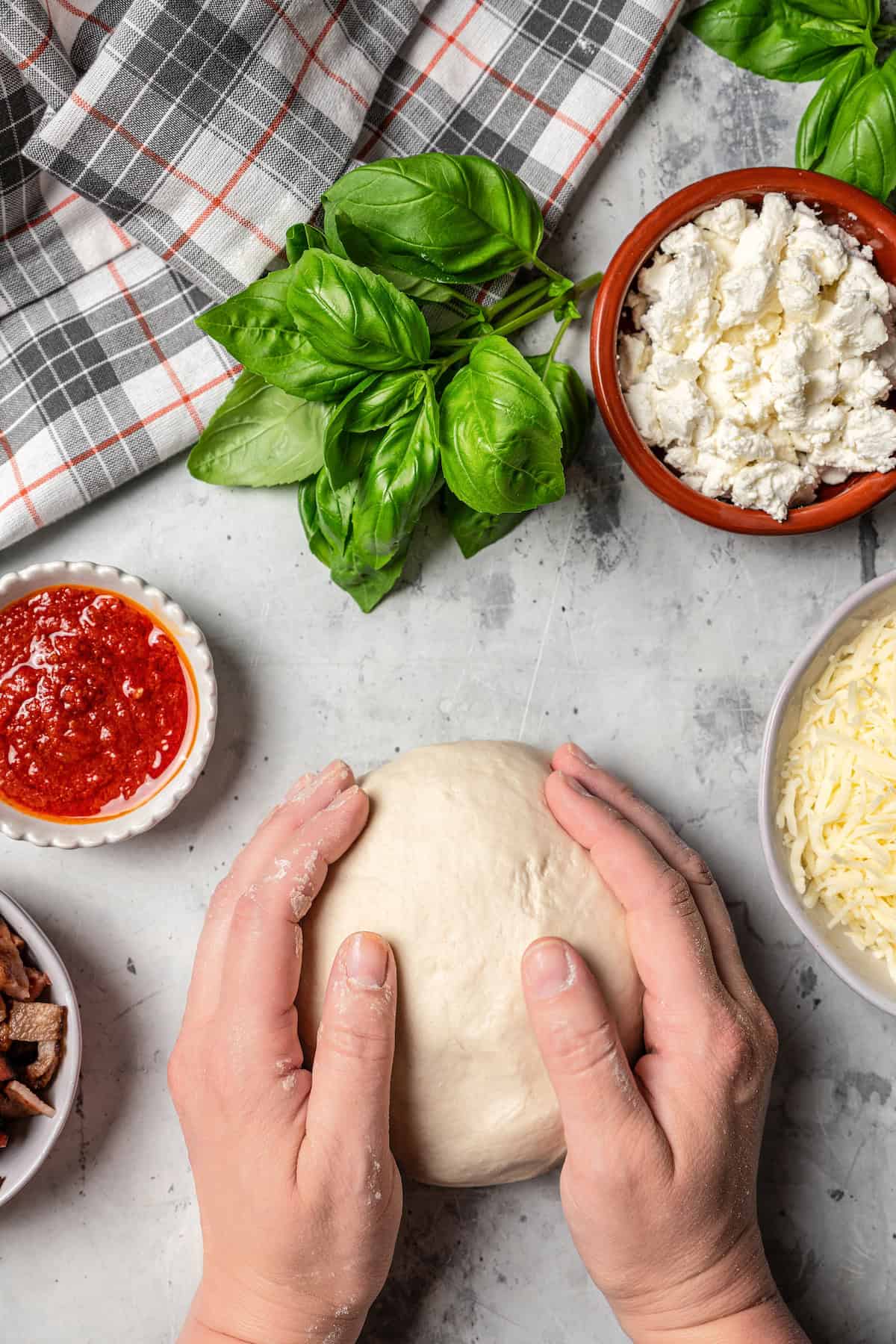 Two hands shaping a ball of pizza dough, surrounded by bowls of pizza toppings.