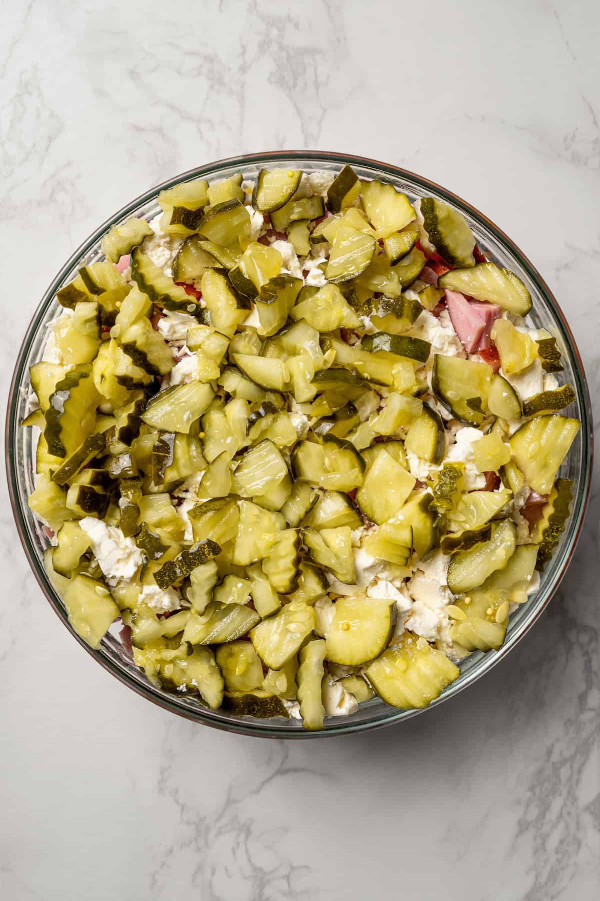 Chopped pickles layered over the other Russian salad ingredients in a glass bowl.