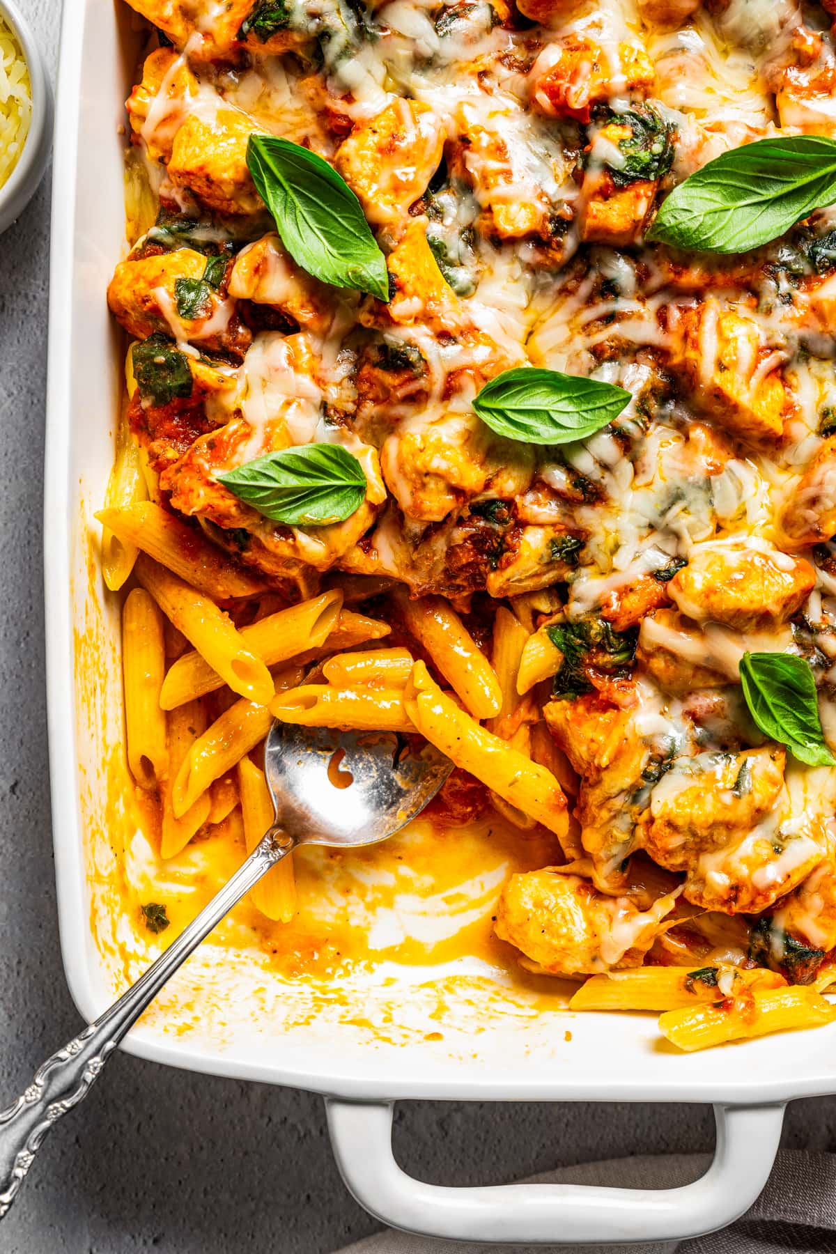 Overhead view of a chicken pasta bake in a casserole dish garnished with basil leaves, with a serving missing from the corner of the pan.