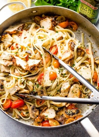 Pair of tongs tossing chicken and pasta with cherry tomatoes and mushrooms in a skillet.