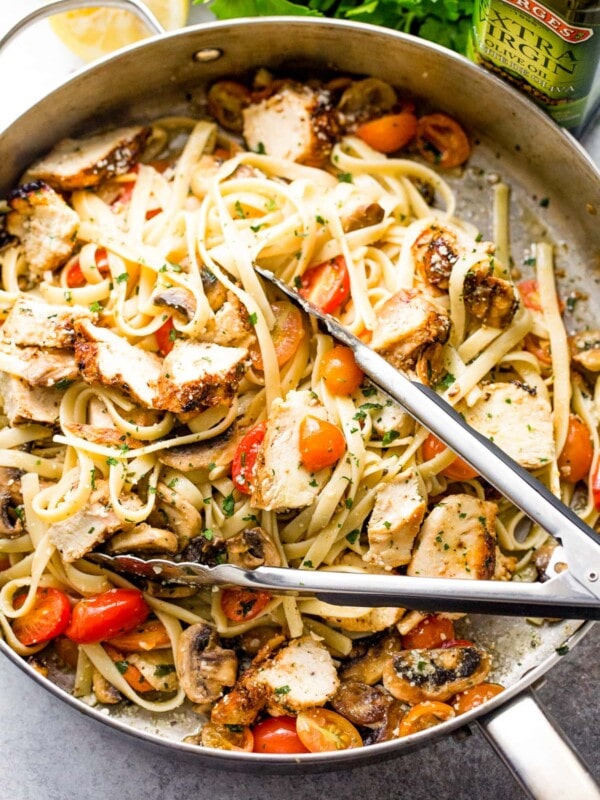 Pair of tongs tossing chicken and pasta with cherry tomatoes and mushrooms in a skillet.