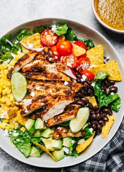 A large bowl with a salad featuring sliced chicken breast, cherry tomatoes, nacho chips, avocado, onions, black beans, and corn.