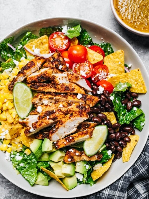 A large bowl with a salad featuring sliced chicken breast, cherry tomatoes, nacho chips, avocado, onions, black beans, and corn.