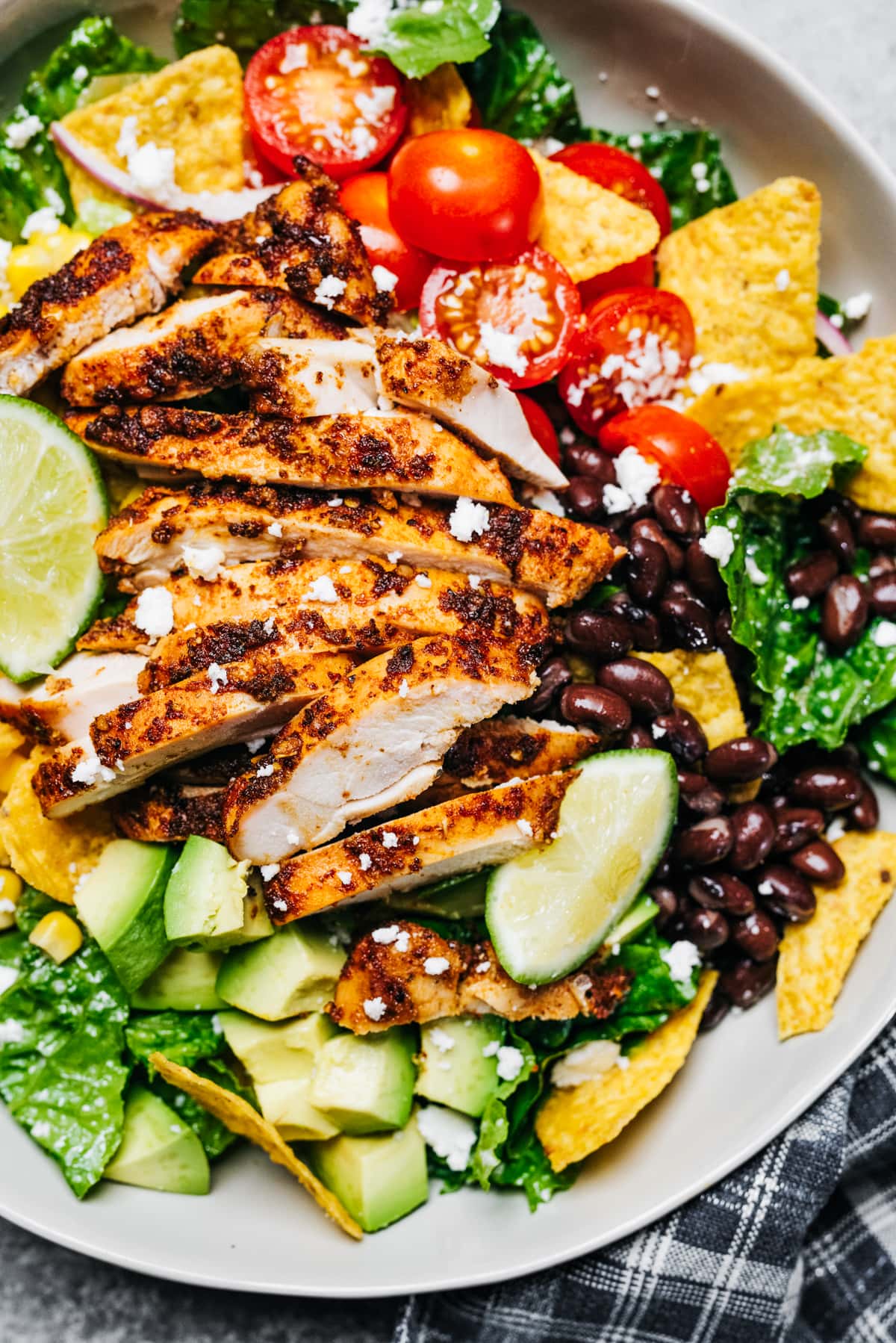 Close-up image of sliced grilled chicken arranged on over lettuce greens, tomatoes, black beans, and nacho chips.