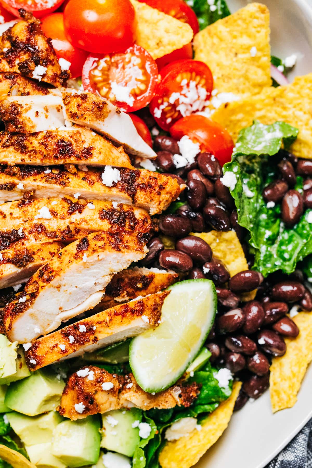 Close-up image of sliced grilled chicken arranged on over lettuce greens, tomatoes, black beans, and nacho chips.
