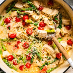 Close up of creamy chicken orzo with asparagus and tomatoes in a large pot with a spoon.