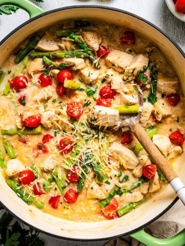Close up of creamy chicken orzo with asparagus and tomatoes in a large pot with a spoon.