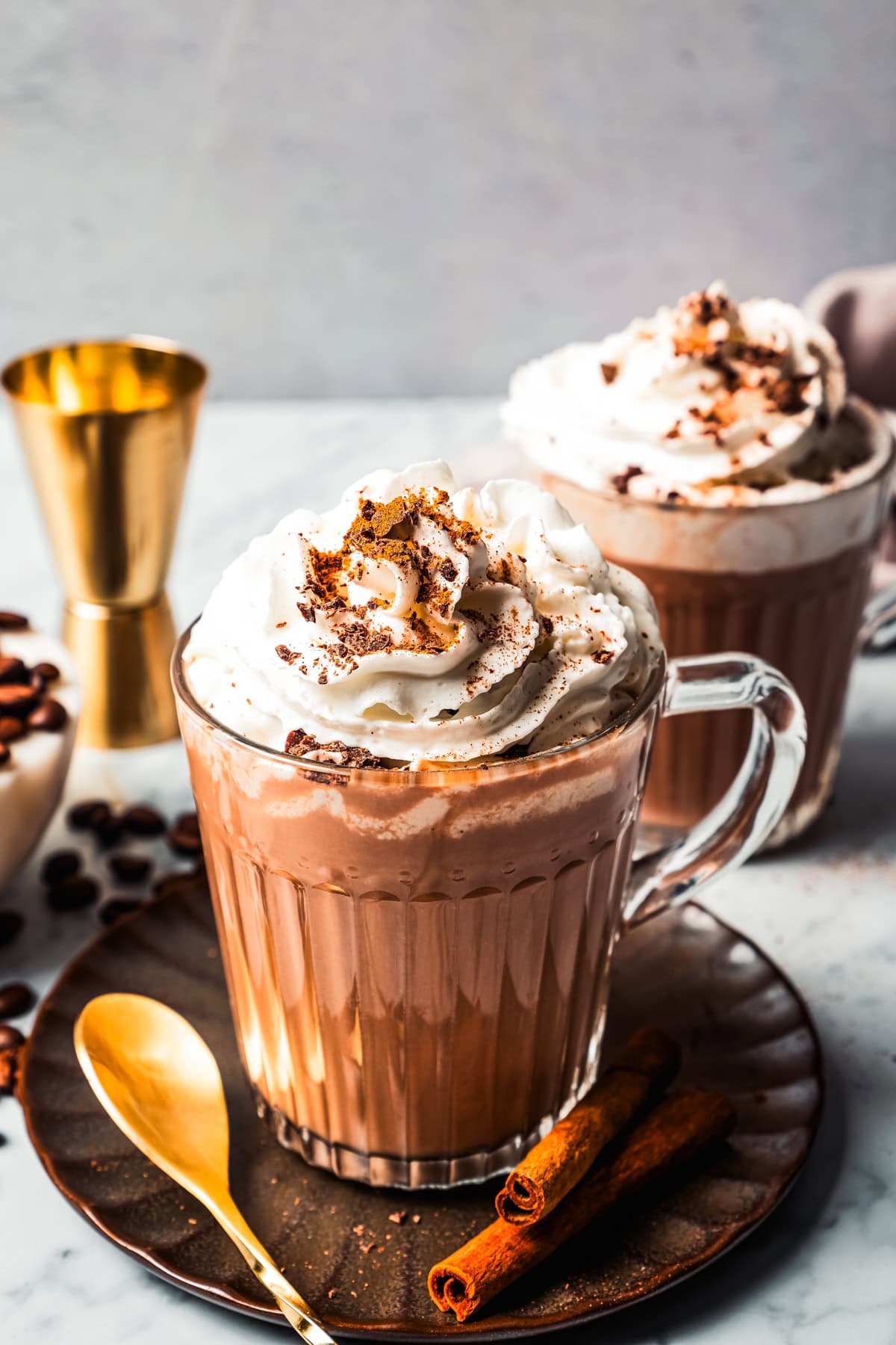 Mexican Coffee Coffee drink topped with whipped cream in a glass mug on a grey stoneware saucer, with a second mug and a cocktail jigger in the background.