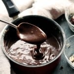 A spoon holding up homemade hot fudge sauce over a pan of more hot fudge sauce.