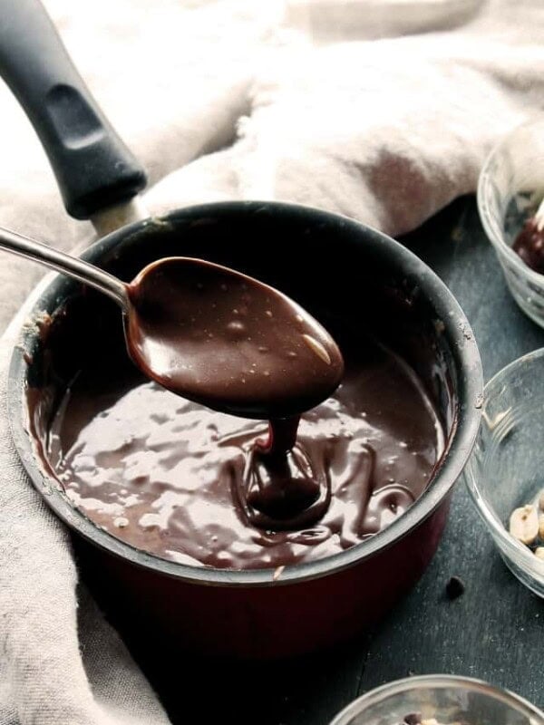 A spoon holding up homemade hot fudge sauce over a pan of more hot fudge sauce.