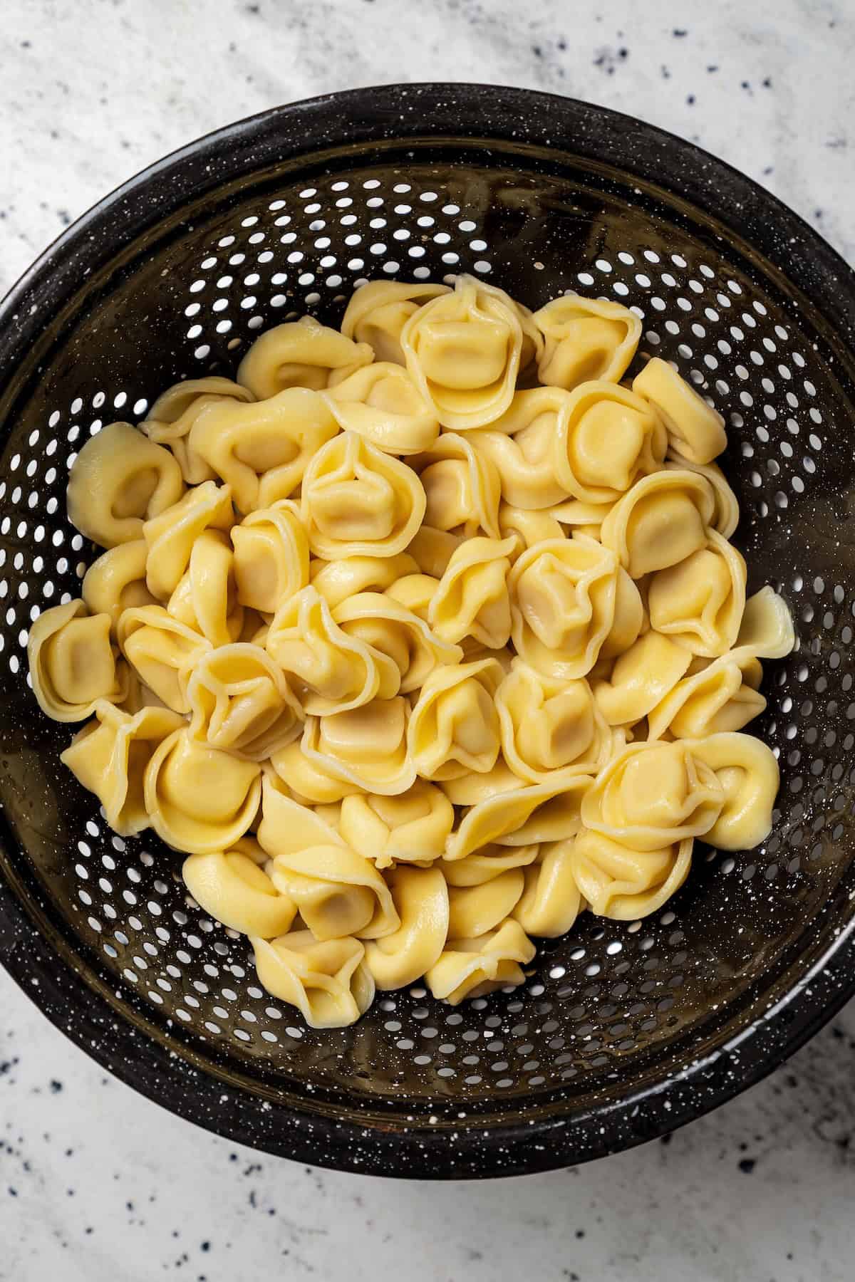 This Mediterranean tortellini pasta salad is a flavorful, easy summer side dish with cheesy tortellini, olives, red peppers, and feta cheese! Cooked and drained tortellini in a black colander.