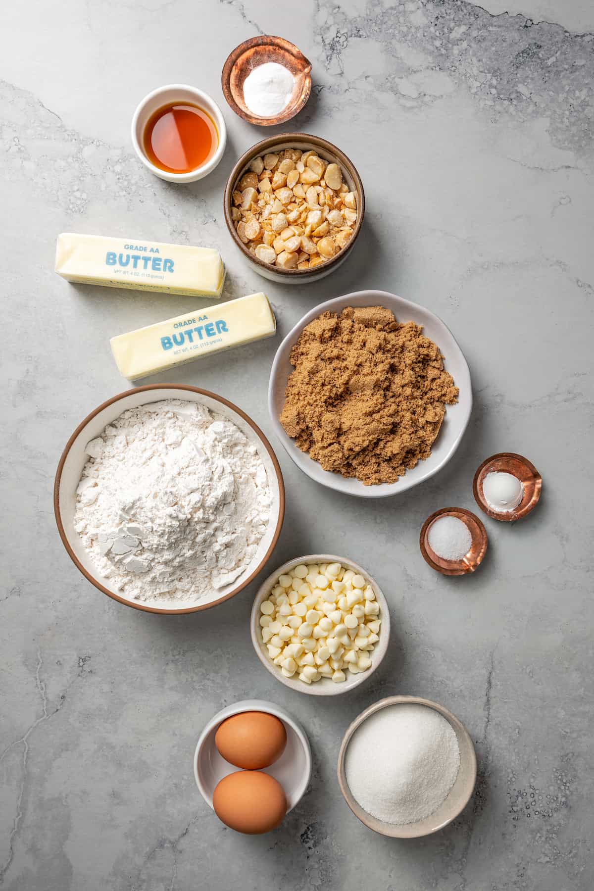 Overhead view of the ingredients needed for white chocolate chip cookies: a bowl of flour, a bowl of white chocolate chips, a bowl of macadamia nuts, a bowl of sugar, a bowl of brown sugar, a bowl of eggs, a bowl of salt, a bowl of baking soda, a bowl of baking powder, a bowl of vanilla extract, and two sticks of butter