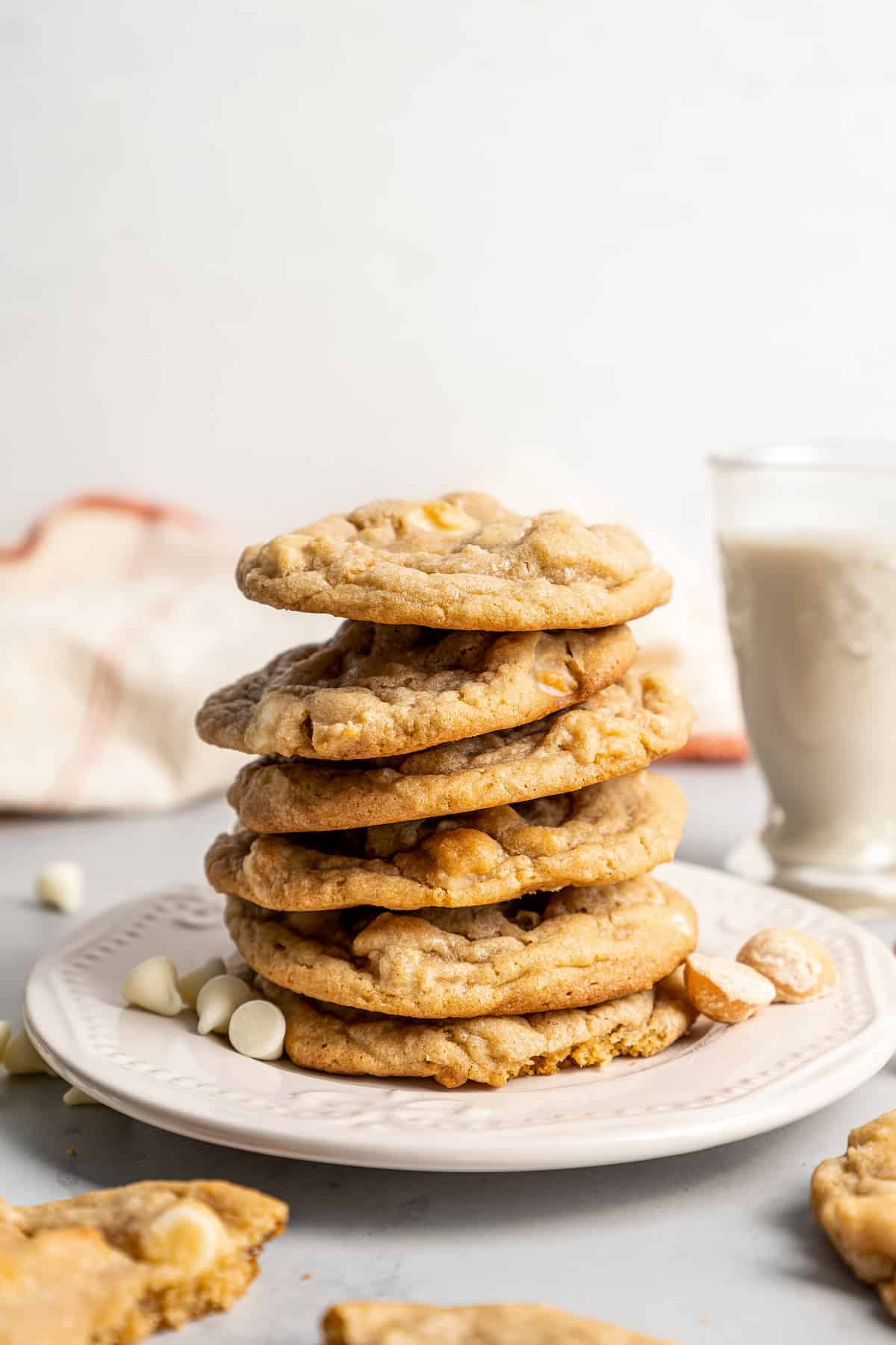 A plate with a stack of six white chocolate chip cookies, surrounded by white chocolate chips, with a glass of milk in the background