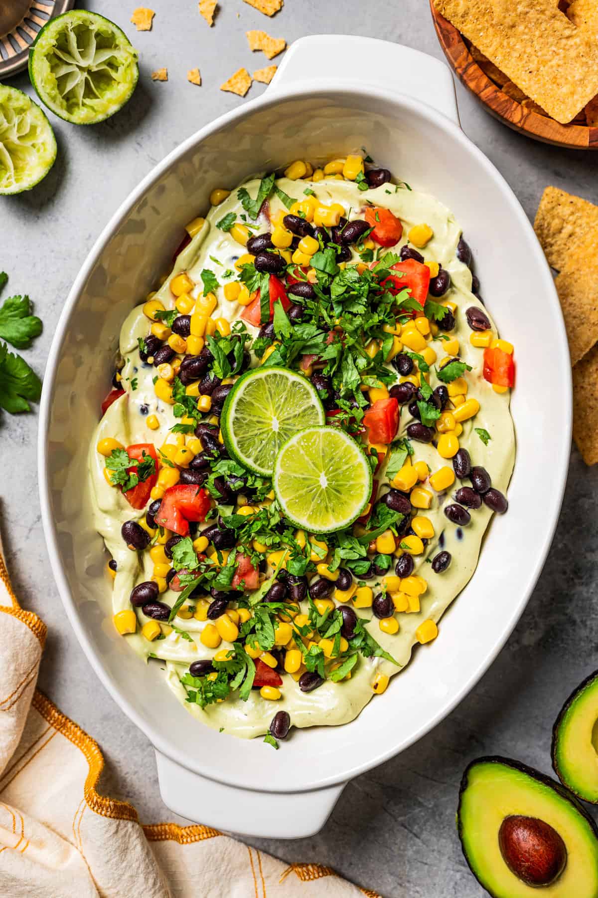 This Southwest avocado dip layers creamy avocado crema with a zesty mix of black beans, sweet corn, and tomatoes. Great with chips or veggies! Overhead view of avocado dip topped with lime wedges and fresh cilantro in a serving dish.