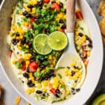 Layered avocado dip topped with lime wedges and fresh cilantro in a serving dish, with a spoon.