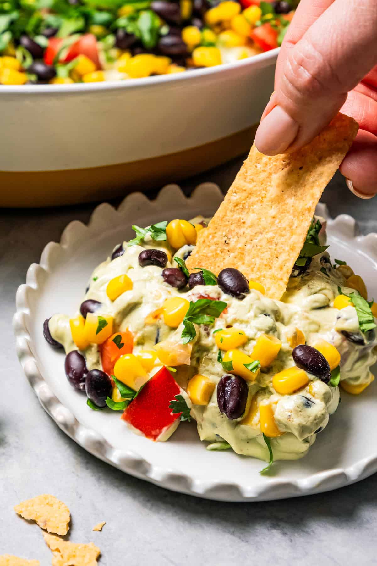 This Southwest avocado dip layers creamy avocado crema with a zesty mix of black beans, sweet corn, and tomatoes. Great with chips or veggies! A hand dipping a tortilla chip into a serving of layered avocado dip on a plate, with the rest of the dip in a serving dish in the background.