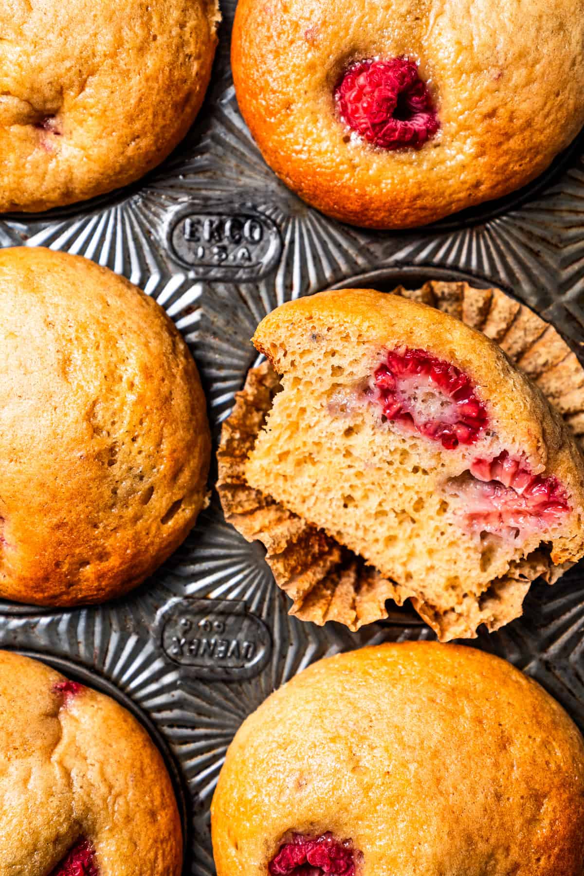 These easy raspberry muffins are fluffy and moist, made with yogurt, brown sugar, and heaps of fresh raspberries. Like summer in a muffin pan! Close up pverhead view of raspberry muffins in a muffin pan, with one muffin cut in half.
