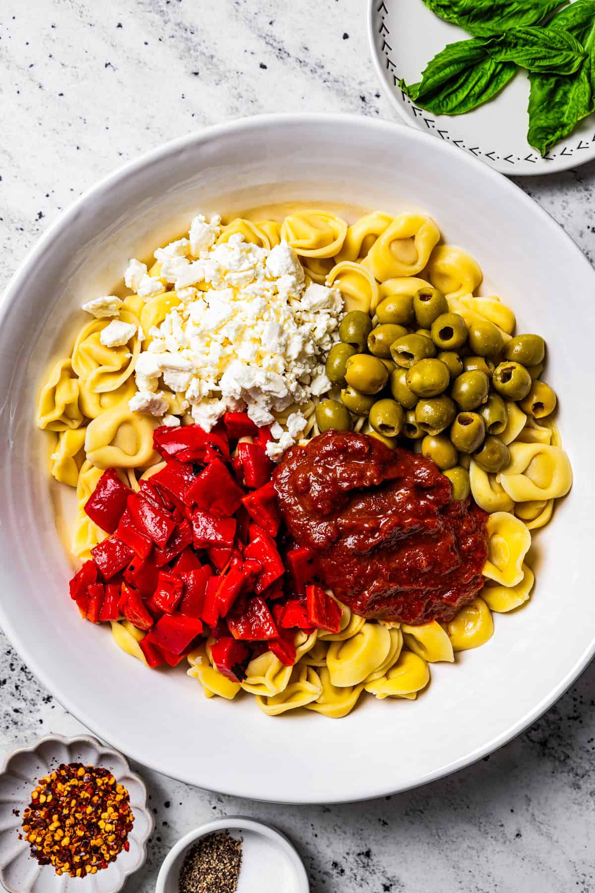This Mediterranean tortellini pasta salad is a flavorful, easy summer side dish with cheesy tortellini, olives, red peppers, and feta cheese! A large bowl with cooked tortellini, olives, tomato sauce, peppers, and cheese.