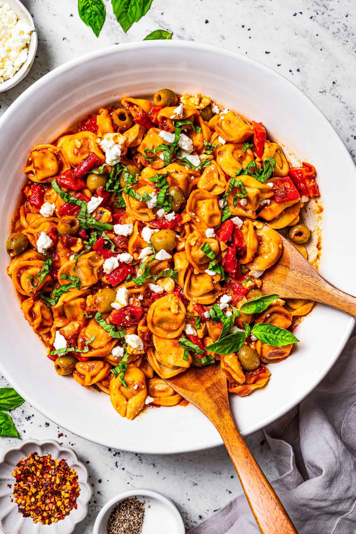 This Mediterranean tortellini pasta salad is a flavorful, easy summer side dish with cheesy tortellini, olives, red peppers, and feta cheese! Tortellini pasta salad in a salad bowl with two wooden spoons placed in the salad.